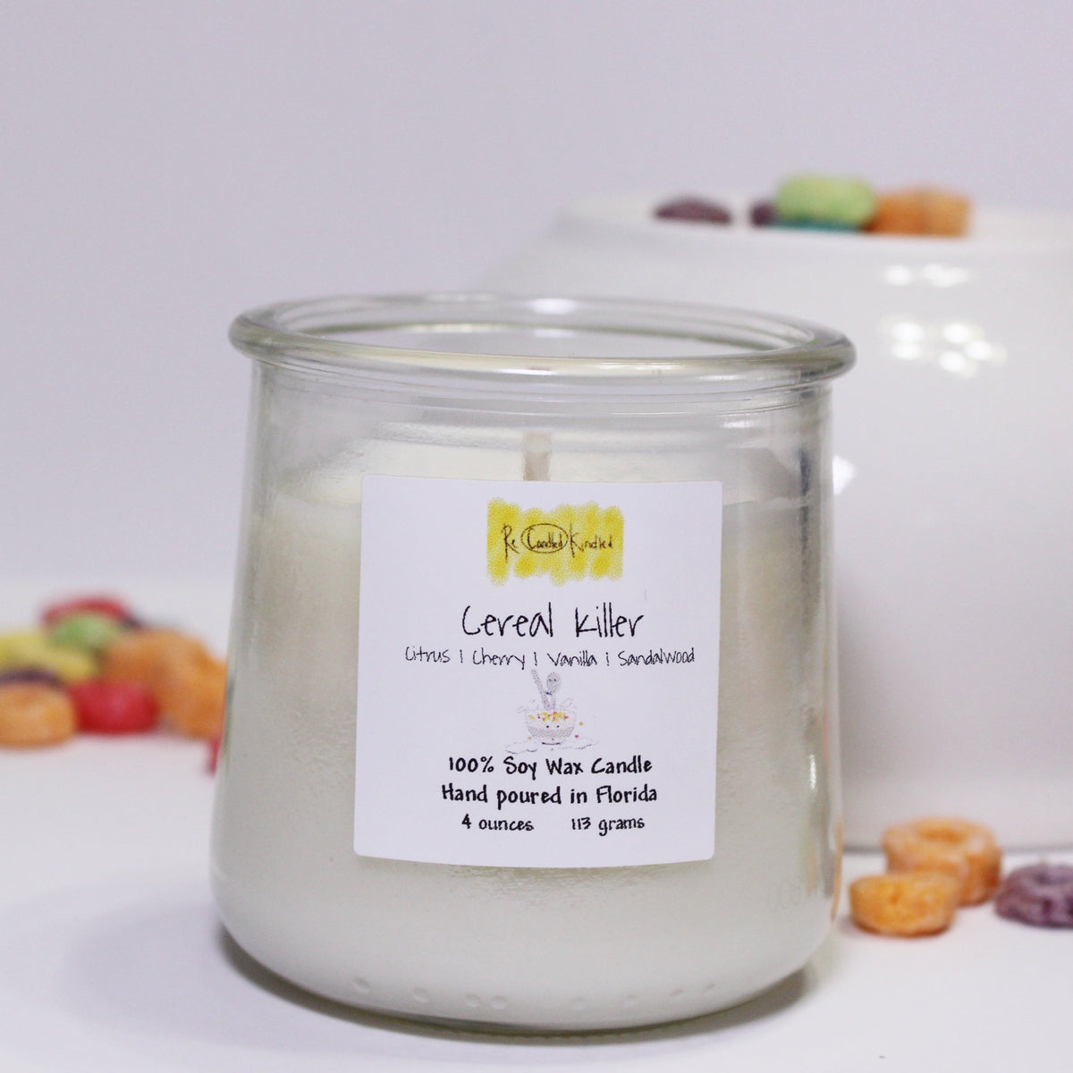 A clear glass candle with a label reading 'CEREAL KILLER' placed on a white surface, surrounded by various colorful cereal pieces.